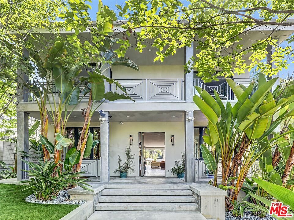 Front facade approached through a tropical entry path with banana palms and bird-of-paradise framing white columns and a wrought-iron balcony.