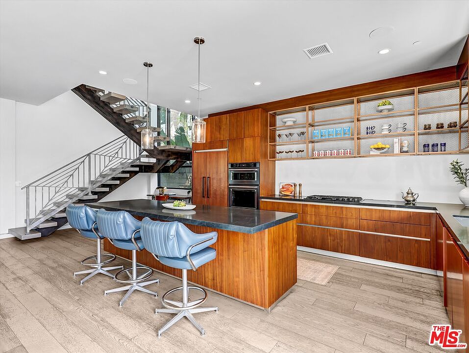 The mahogany kitchen island with blue leather stools, anchored by the floating steel staircase rising into the second floor.