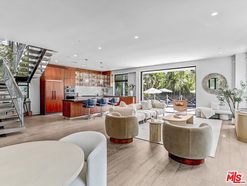 The great room at 1724 N Sierra Bonita, with a floating staircase, mahogany and white kitchen, and a wall of glass retracted onto the pool deck and tropical garden.